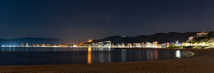 The panoramic view of a coastal town at night with reflections in the water. Cullera, Spain.