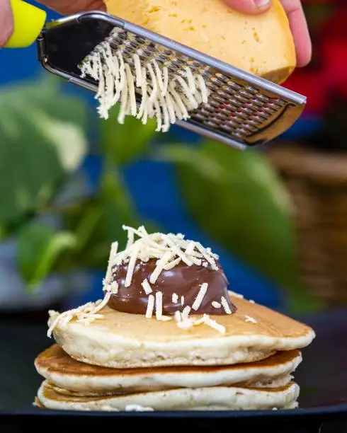 A vertical shot of a gouda cheese being grated on hotcakes with chocolate on top