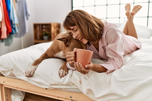 Young caucasian woman drinking coffee lying on bed with dog at bedroom