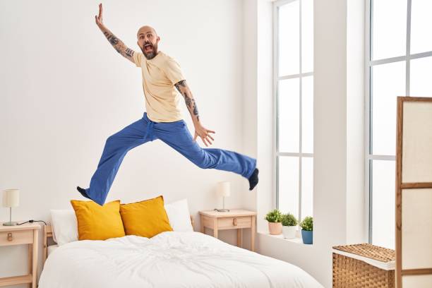 Young bald man jumping on bed at bedroom