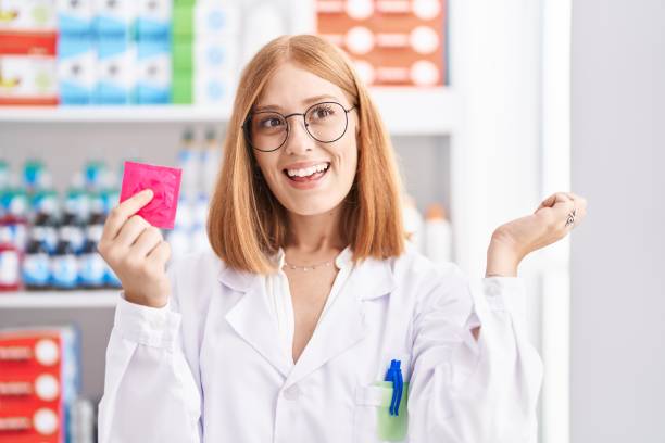 молодая рыжеволосая женщина, работающая в аптеке аптеки, держащая презерватив с гордым криком, празднующая победу и успех, очень взволнова� - care condom confidence contraceptive стоковые фото и изображения