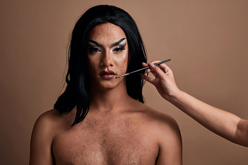 Makeup, beauty and lgbtq male model and hand of a stylist in studio on a wall background. Cosmetics, empowerment and inclusion with a male drag queen backstage with a beautician