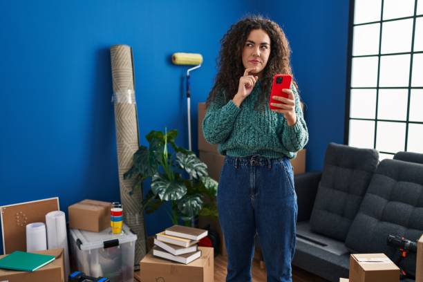 istock Young hispanic woman at new home using smartphone serious face thinking about question with hand on chin, thoughtful about confusing idea 1437881824