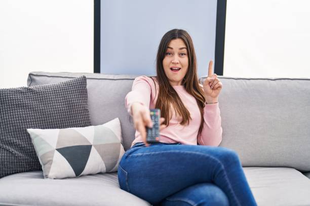 istock Young brunette woman holding television remote control surprised with an idea or question pointing finger with happy face, number one 1437884876