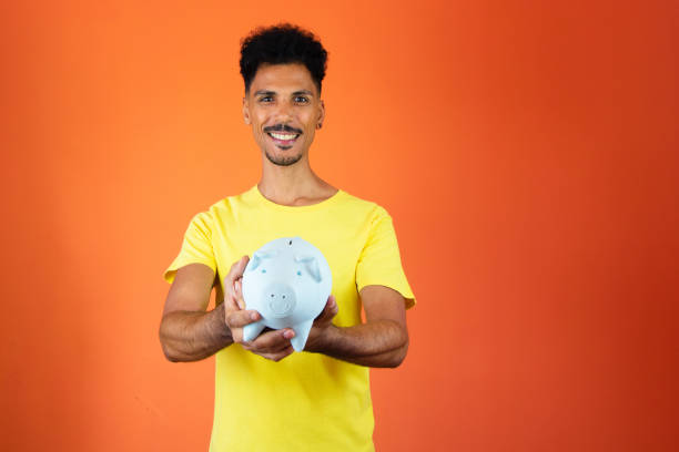 Handsome Black Man Holding a Piggy Bank Isolated on Orange. Man in a yellow shirt isolated. stock photo