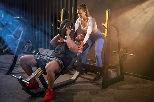 Muscular young man lifting heavy weight plate with assistance of attractive young fit woman at health club gym.