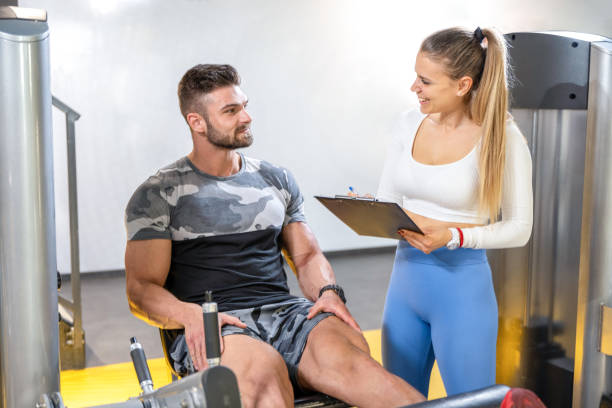 istock Muscular handsome man doing exercises on leg press machine and female personal trainer with clipboard calculating his muscle progress during workout class at gym. 1437887633