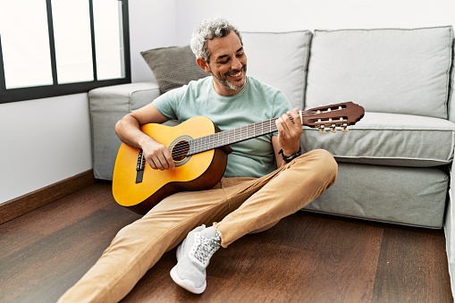 Middle age grey-haired man playing classical guitar sitting on floor at home