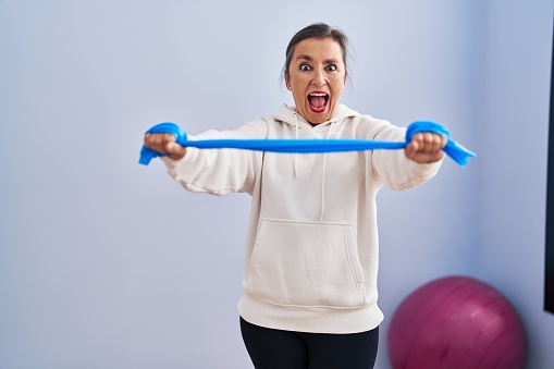 Middle age hispanic woman training arm resistance with elastic arm bands celebrating crazy and amazed for success with open eyes screaming excited.