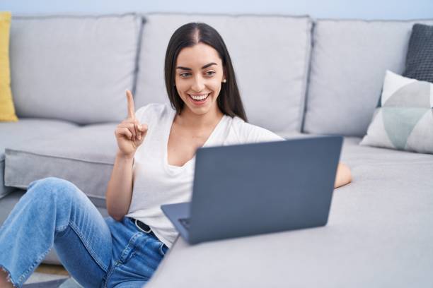 istock Young brunette woman using laptop at home smiling with an idea or question pointing finger with happy face, number one 1437895099