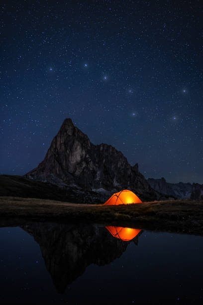 Awesome night sky with many stars and the constellation of the big dipper above Mountain peak in the dolomites and illuminatet tent at lake, everything reflecting in dark water