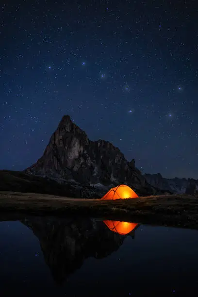 Awesome night sky with many stars and the constellation of the big dipper above Mountain peak in the dolomites and illuminatet tent at lake, everything reflecting in dark water
