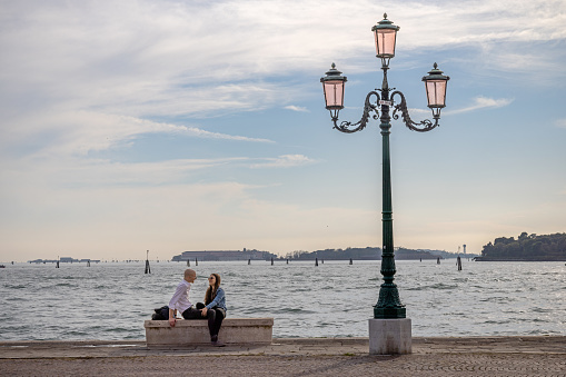 Venice, Italy - October 11th 2022: Couple sitting on a bench under a street lamp on the waterfront in the center of the old and famous Italian city Venice