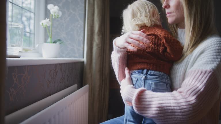 Mother Cuddling Son Trying To Keep Warm By Radiator At Home During Cost Of Living Energy Crisis