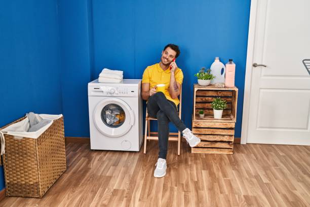 istock Young hispanic man drinking coffee talking on the smartphone wating for washing machine at laundry room 1437899825