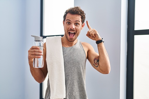 Young hispanic man wearing sportswear drinking water smiling amazed and surprised and pointing up with fingers and raised arms.