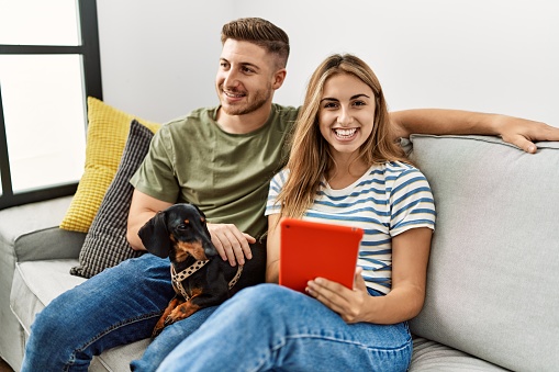 Young hispanic couple smiling happy using touchpad sitting on the sofa with dog at home.
