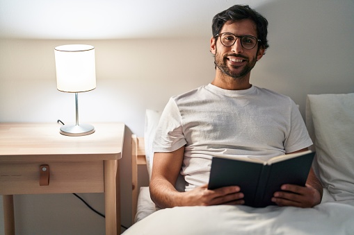 Young hispanic man reading book sitting on bed at bedroom