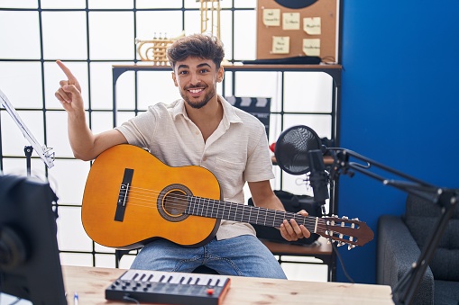 Arab man with beard playing classic guitar at music studio smiling happy pointing with hand and finger to the side