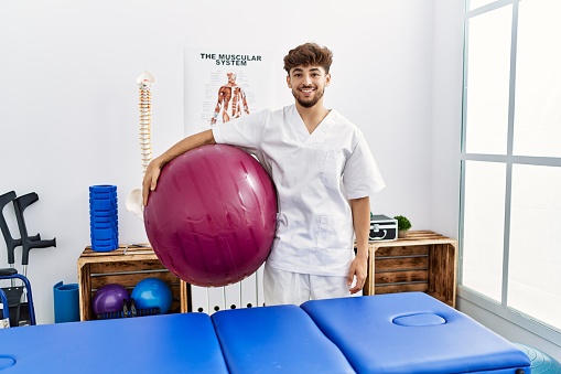 Young arab man wearing physiotherapist uniform holding fit ball at clinic