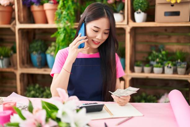 フラワーショップでスマートフォンで話すドルを数える若い中国人女性の花屋 - flower shop flower flower market store ストックフォトと画像