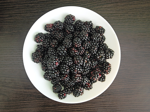 Black juicy blackberries on a white plate on a dark wooden background.