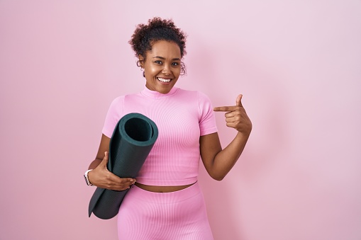 Young hispanic woman with curly hair holding yoga mat over pink background looking confident with smile on face, pointing oneself with fingers proud and happy.