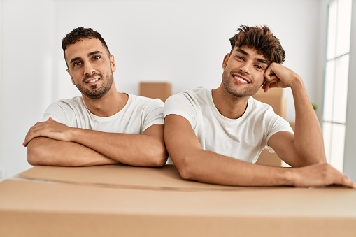 Two hispanic men couple smiling confident leaning on cardboard box at new home