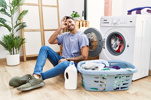 Young hispanic man putting dirty laundry into washing machine doing ok gesture with hand smiling, eye looking through fingers with happy face.