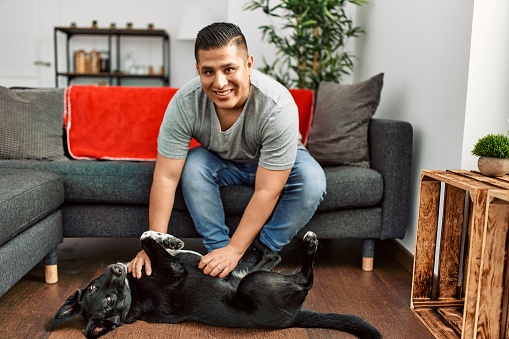 Young hispanic man smiling happy playing with dog at home.