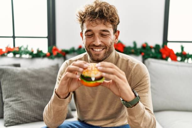 Young hispanic man eating hamburger sitting by christmas decor at home stock photo