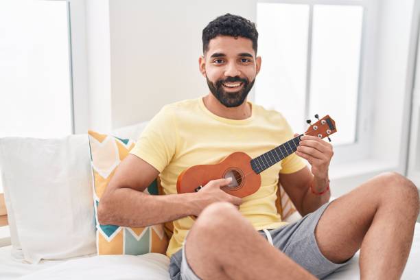 istock Young arab man playing ukulele sitting on bed at bedroom 1437917543