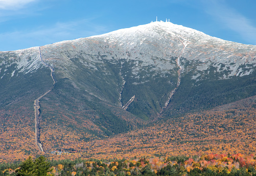 Blue sky and colorful fall foliage with dusting of snow atop Mount Washington in New Hampshire. Visible on left are tracks of cog railway and antenna of weather observatory on summit.