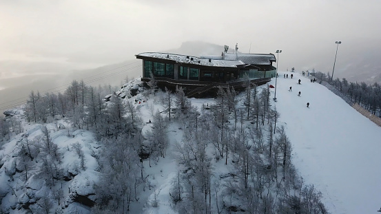 Aerial view of the ski resort with snowy mountain slopes and winter trees. Stock footage. Upper station of cableway and a group of people going skiing and snowboarding down the hill.
