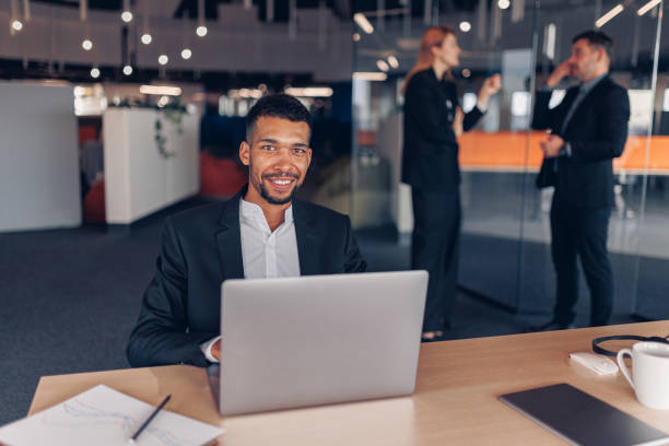istock Smiling african businessman looking at camera while working laptop in modern office 1437923128