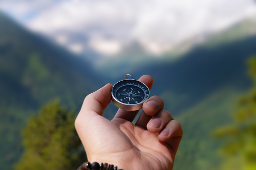 A hand with a compass against the backdrop of epic snow-capped mountains with clouds and a forest at the foot, close-up.