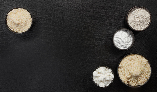 Black bowls with different types of flour on a black slate board, top view. Space for text.