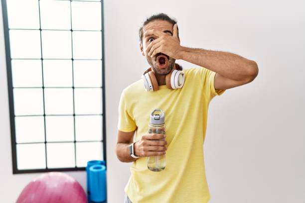 joven hispano vestido con ropa deportiva y bebiendo agua en el gimnasio mirando en estado de shock cubriéndose la cara y los ojos con la mano, mirando a través de los dedos con expresión avergonzada. - hiding fear men peeking fotografías e imágenes de stock