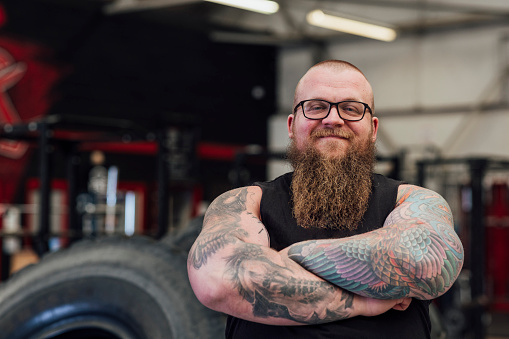 A strongman standing next to a truck tyre in a gym in Newcastle upon Tyne, England. He is looking at the camera and smiling with his arms crossed.