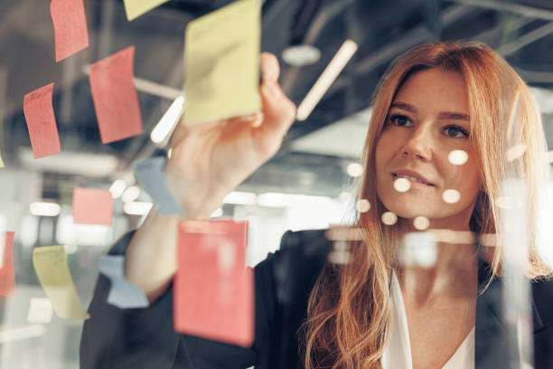 Businesswoman writing on sticky notes on glass wall while working in office stock photo