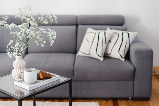 vase with flowers, fresh croissant and cup of coffee on marble tray on glass side table, comfort couch with pillows in lounge room at apartment with scandinavian interior