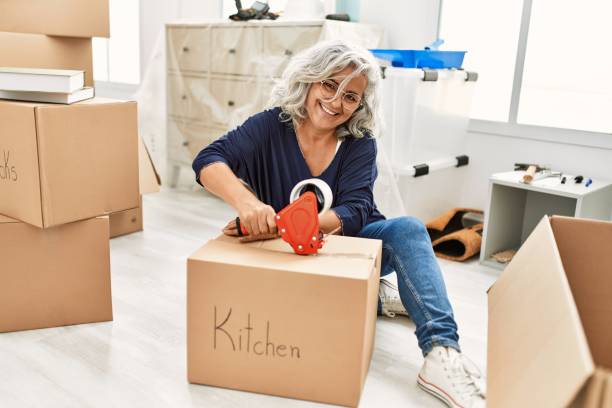 Middle age grey-haired woman smiling happy packing kitchen cardboard box at new home. Middle age grey-haired woman smiling happy packing kitchen cardboard box at new home. grey hair on floor stock pictures, royalty-free photos & images