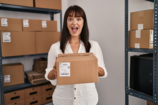 Young brunette woman working at small business ecommerce holding package celebrating crazy and amazed for success with open eyes screaming excited.