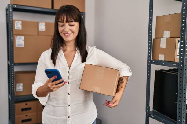istock Young beautiful hispanic woman ecommerce business worker using smartphone holding package at office 1437929334