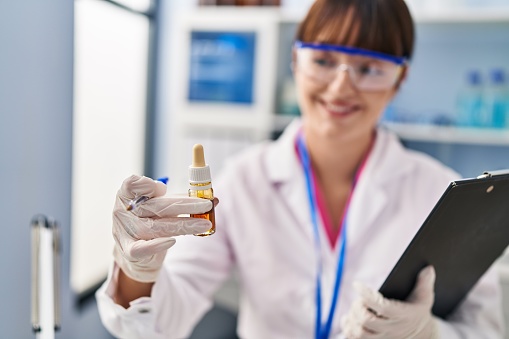 Young beautiful hispanic woman scientist holding clipboard and bottle at pharmacy