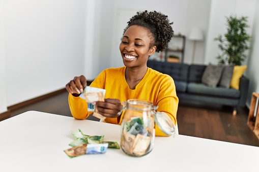 African american woman sitting on table holding rand banknotes of charity jar at home