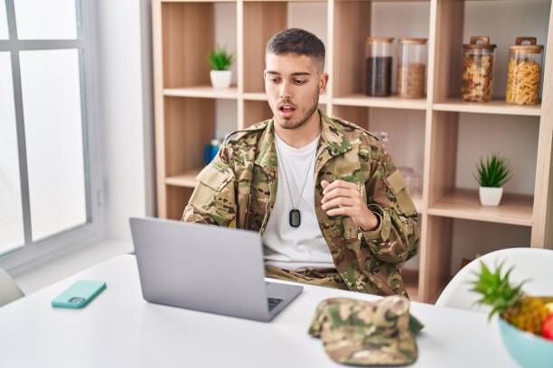 istock Young hispanic man wearing camouflage army uniform doing video call scared and amazed with open mouth for surprise, disbelief face 1437931700