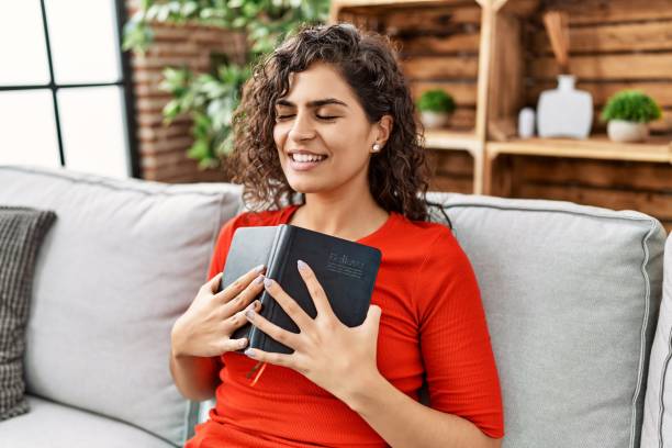 joven latina sonriendo confiada abrazando la biblia en casa - christian mujer leyendo la biblia fotografías e imágenes de stock