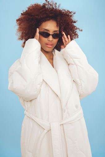 Young woman standing wrapped in a big white winter jacket wearing cool sunglasses. Shot in studio against a blue background