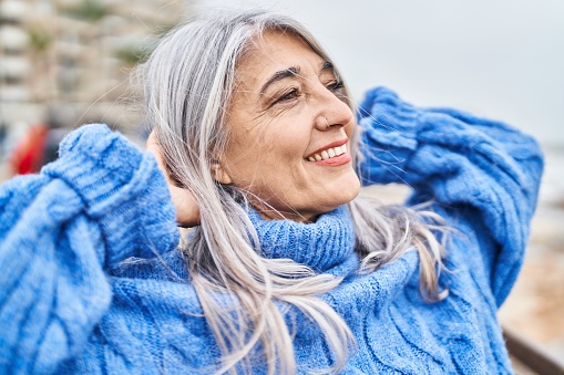 Mujer de cabello gris de mediana edad sonriendo confiada relajada con las manos en la cabeza a la orilla del mar photo